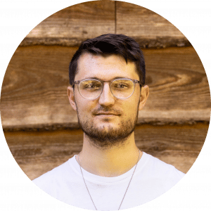 Portrait of a young man with dark hair and glasses against a wooden background.
