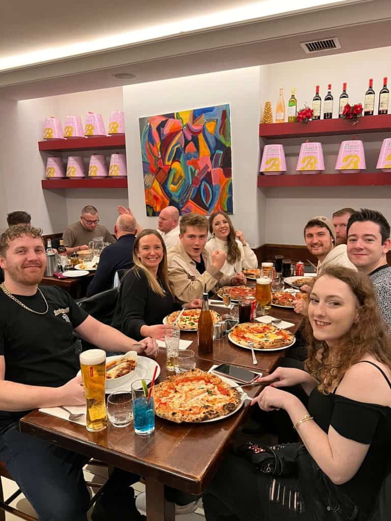People enjoying a dinner with pizza and drinks at a restaurant, smiling and having a good time, vibrant atmosphere, colourful decor on the wall, relaxed social gathering, contemporary indoor setting, casual attire, lively mood, group of friends, dining scene.