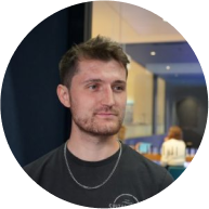 Young man with short brown hair and a beard, wearing a black T-shirt and silver chain, standing indoors at an event or conference, with a blurred background and a blue ceiling light.