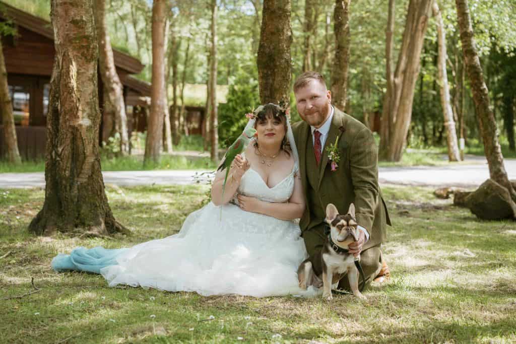 Flower crown-wearing bride and groom with a dog and bird during outdoor wedding in forest setting.